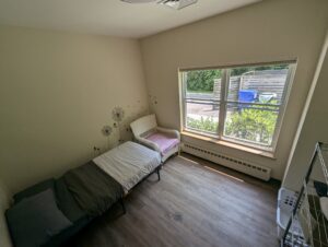 A private bedroom in Howard Center’s Adult Crisis Stabilization program, featuring a single bed with white and gray bedding, a wicker chair, a large window overlooking a garden, and minimalist wall decor with dandelion designs. The room is softly lit, with wooden flooring and a serene atmosphere.