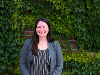 "A woman with long dark hair, wearing a gray blazer over a black and white polka dot top, stands smiling in front of a brick wall covered in green ivy